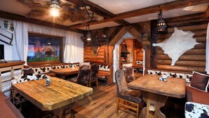 Interior rústico de madera con mesas y sillas de madera, un ventanal con vista a una casa y un trofeo de piel de animal en la pared.