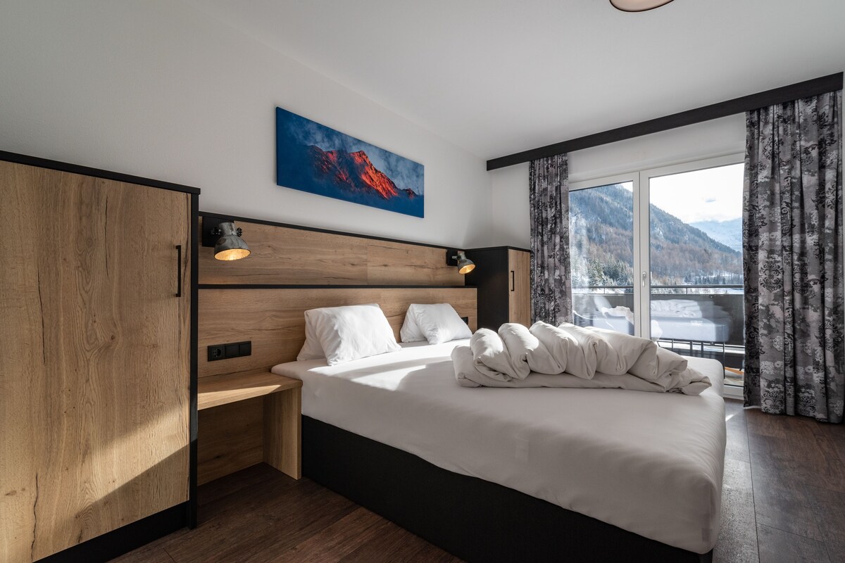 A modern hotel room with a large bed, wooden headboard, a painting of a mountain, and a view of mountains through the window.