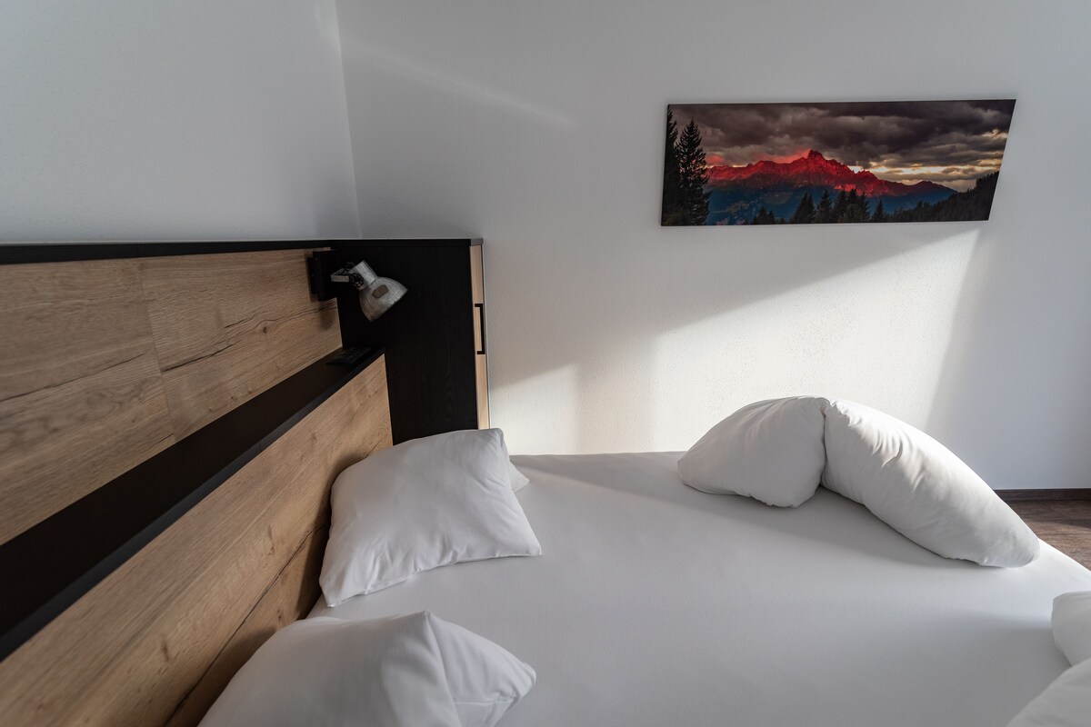 A neatly made bed with white bedding and pillows, a wooden headboard, and a painting of a mountain landscape on the wall.