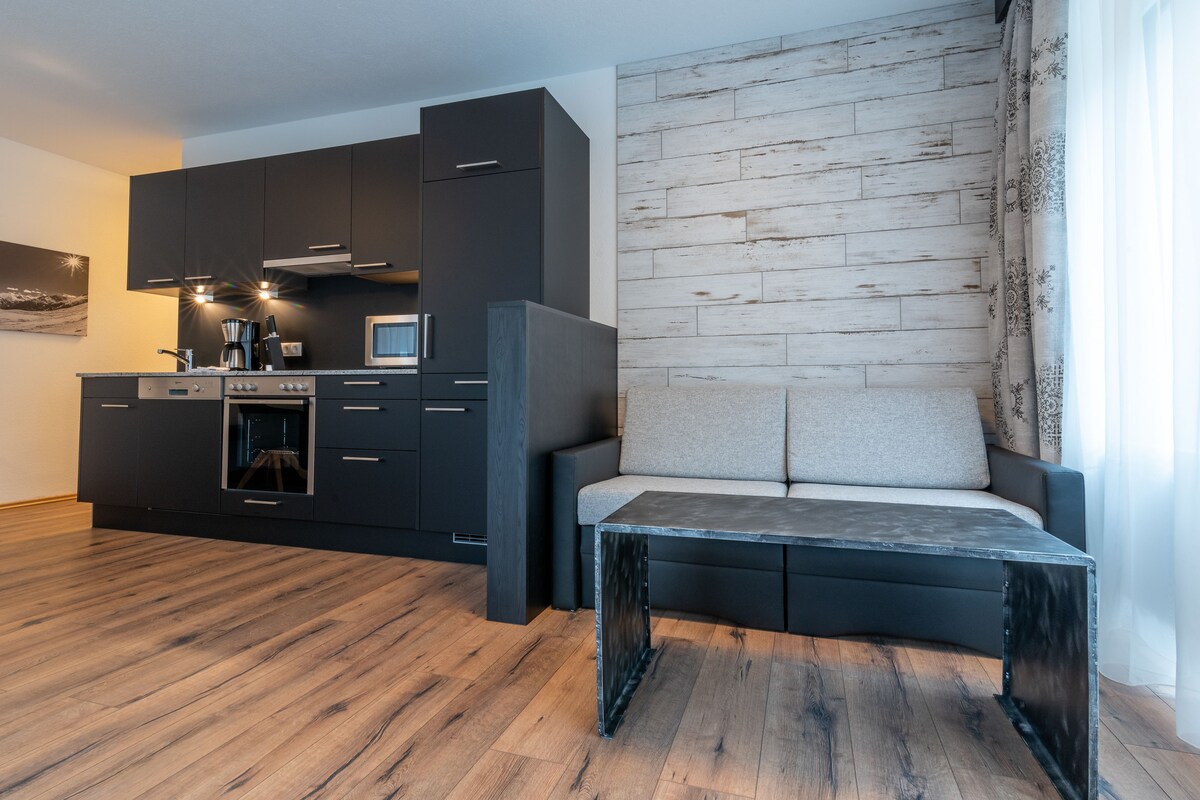 A modern kitchen with dark cabinetry, a built-in oven, and a microwave. There is a wooden floor and a seating area with a cushioned bench.