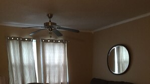 A living room with a wooden floor, a round table with a green tablecloth, a dark-colored sofa, a wooden chair, a ceiling fan, and a round mirror on the wall.