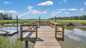Une jetée en bois s’étendant dans une étendue d’eau calme, entourée de prairies verdoyantes et d’arbres sous un ciel dégagé.