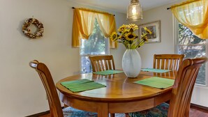 Une salle à manger avec une table en bois, quatre chaises, un vase avec des tournesols, des rideaux jaunes et un luminaire suspendu.