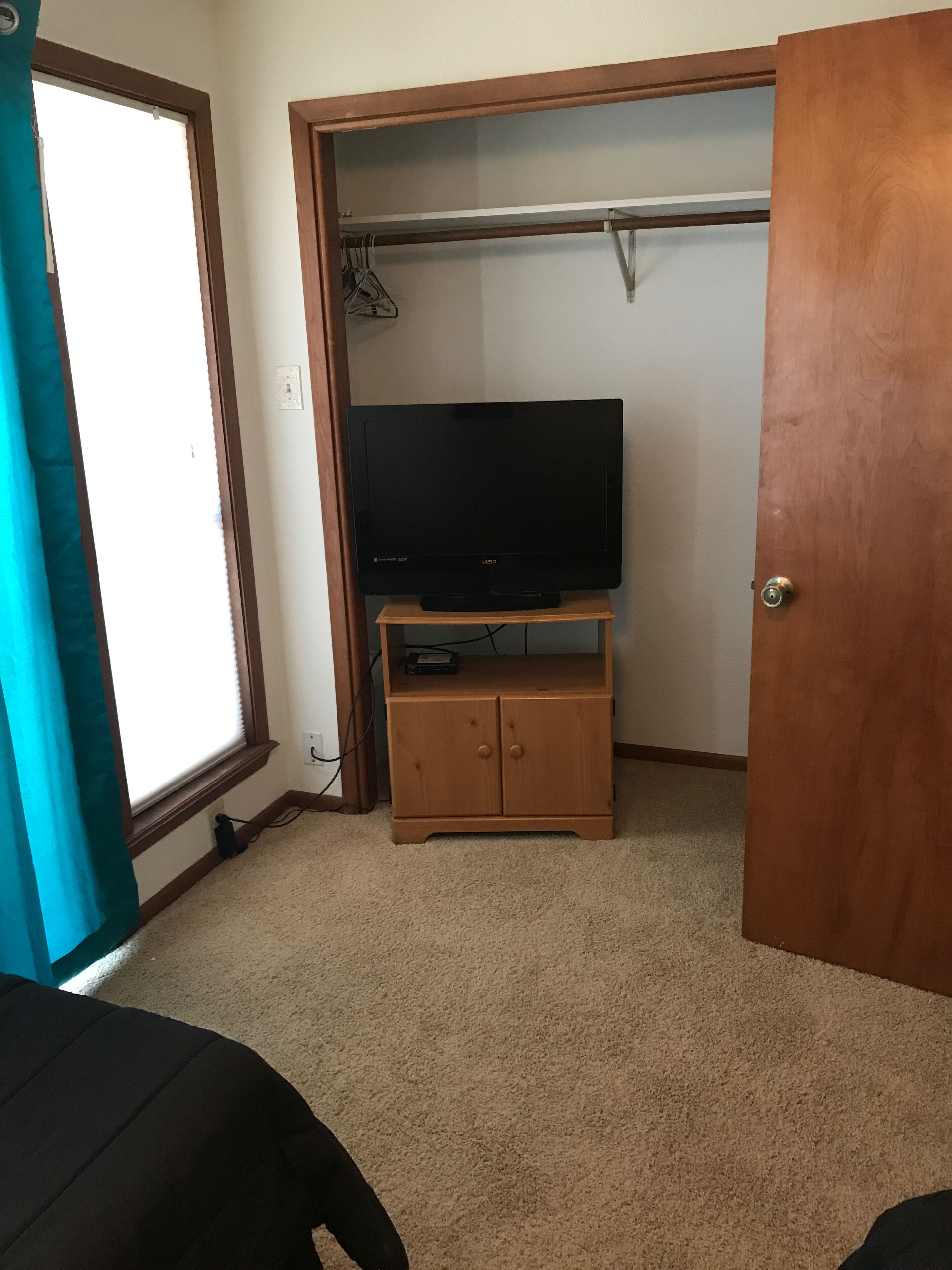 A room with a television on a wooden stand, a closet with a hanging rod, and a window with blinds.