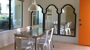 Un coin repas avec une table en verre et des chaises blanches, un luminaire suspendu et un grand miroir au mur.