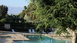 Une piscine entourée de chaises longues et d’une clôture, avec des arbres tout autour et des montagnes en arrière-plan.