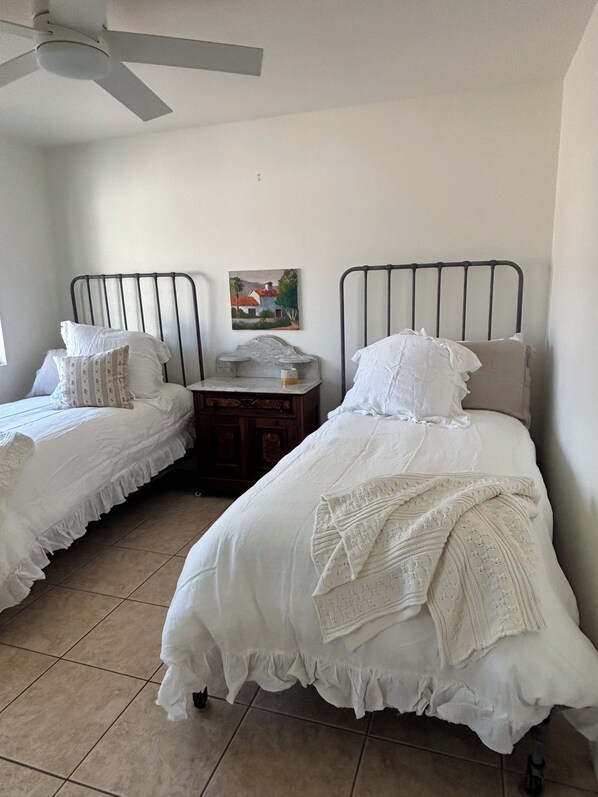 A bedroom with two beds, a ceiling fan, a small table, and a painting on the wall.