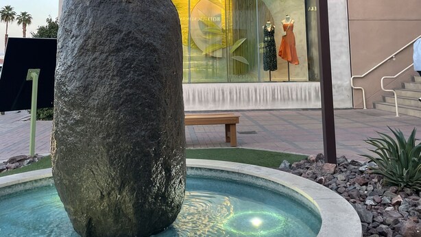Un bâtiment moderne avec un grand élément décoratif en pierre, une piscine et une vitrine ornée de mannequins.