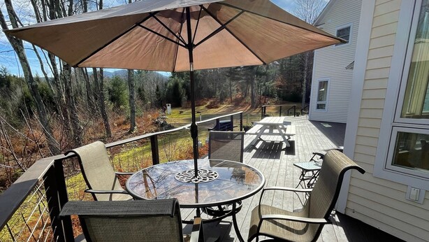 Een houten terras met een ronde tafel en stoelen, een parasol, en uitzicht op een huis en bos.