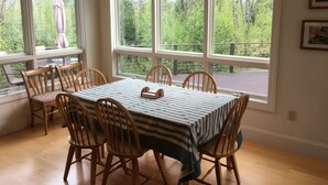 Een eetruimte met een tafel voor zes personen, omringd door houten stoelen, grote ramen met uitzicht op bomen en een weg, en een klein beeldje op de vensterbank.