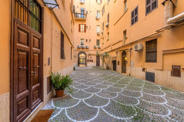 A narrow alleyway with patterned cobblestone pavement, wooden doors, and balconies with air conditioning units.