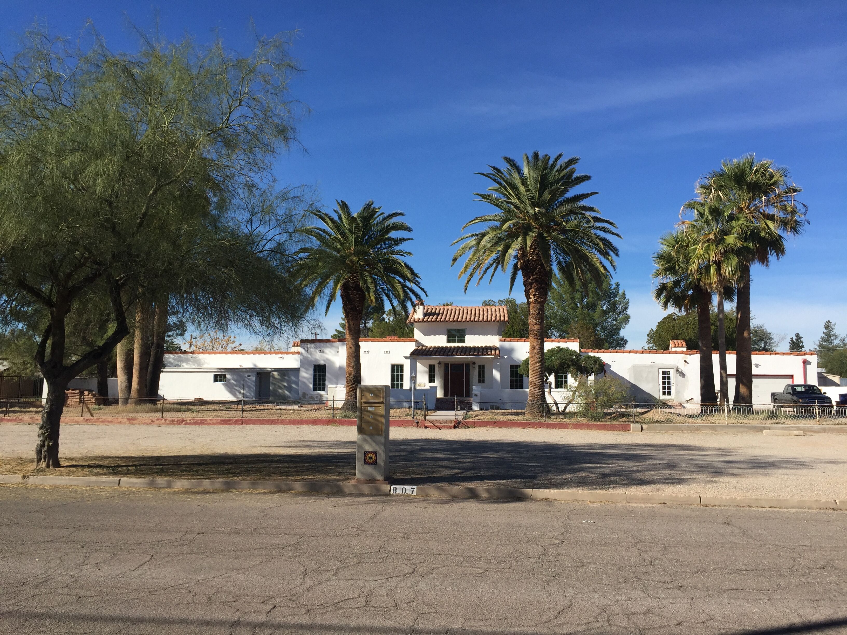 A building with a white facade and red-tiled roof, surrounded by palm trees and a fenced area.