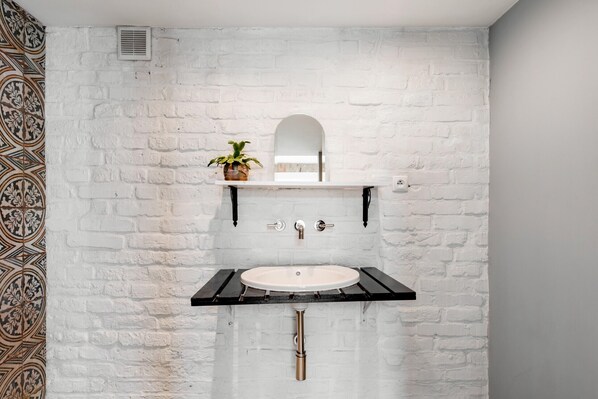 A bathroom with a white brick wall, a white sink with a black countertop, a small shelf with a potted plant, and a mirror above the sink.