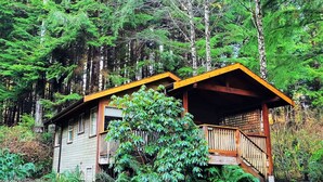Una cabaña con terraza de madera y un porche pequeño, rodeada de un bosque frondoso.