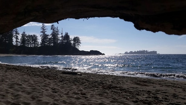 Una playa con la entrada de una cueva, orilla arenosa, olas del océano y una isla boscosa en la distancia.