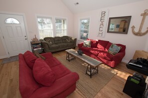 A living room with a red sofa, a green sofa, and a wooden coffee table. There are two windows with blinds, a door, and a wall-mounted anchor decoration.