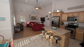 A spacious kitchen with a central island, wooden cabinets, stainless steel appliances, and a red sofa in the living area. - Soundside Oasis: Short Walk to Sound + Linens & Cleaning Included – Sleeps 8 (Kill Devil Hills)