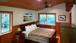 A bedroom with a wooden ceiling, a bed with a patterned bedspread, two windows with views of trees, and a ceiling fan. - Salal Cabin by Evergreen Forest Cabins (Ucluelet)