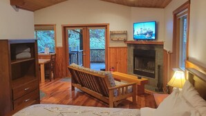 A cozy room with a wooden bench, a fireplace, a television, and a large window overlooking a deck. - Salal Cabin by Evergreen Forest Cabins (Ucluelet)