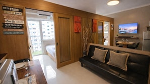 Een moderne woonkamer met een zwarte leren bank, een eethoek met een tafel voor twee personen, en een aan de muur bevestigde televisie.