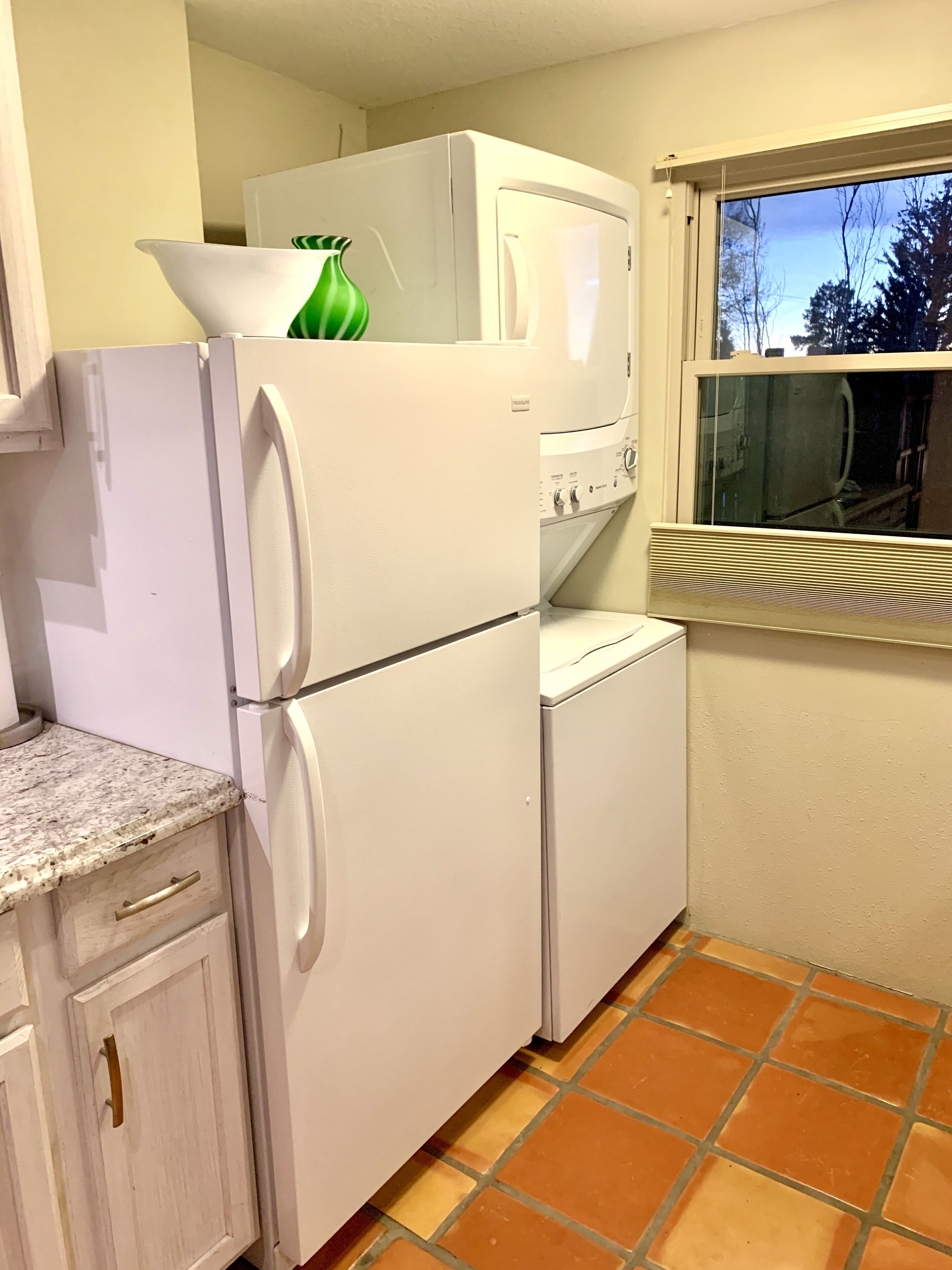 Une cuisine compacte équipée d’un réfrigérateur, d’un lave-linge et d’une fenêtre donnant sur les arbres.