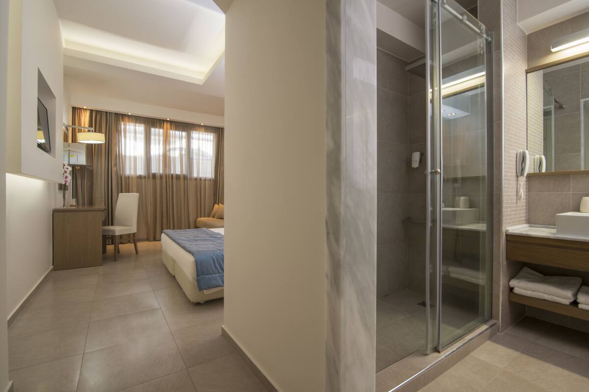 A hotel room with a bed, a desk, a chair, a shower area, and a bathroom with a sink and mirror.