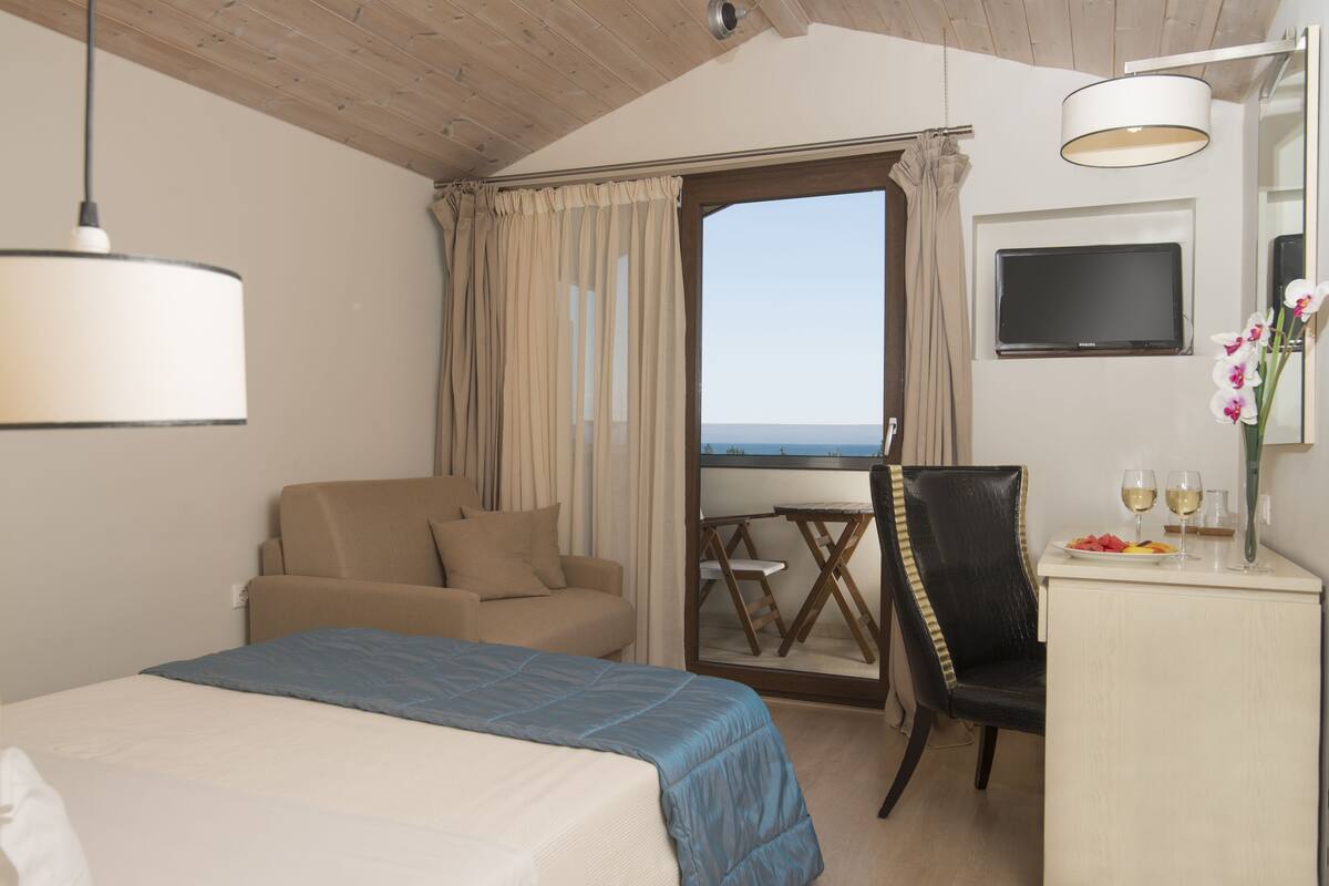 A hotel room with a bed, a chair, a desk with a plate of fruit and two glasses, a TV, and a view of the sea.