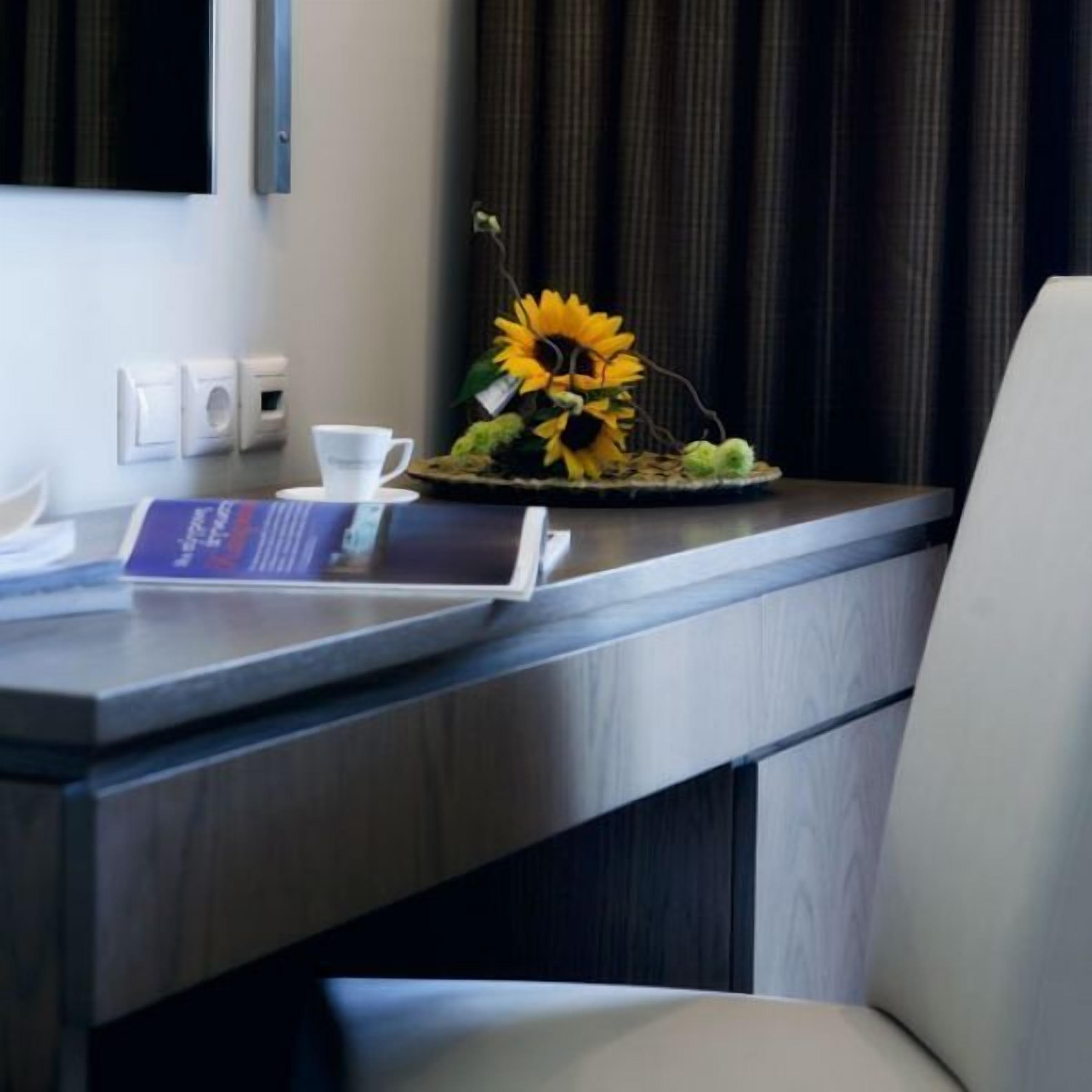 A hotel room with a wooden desk, a laptop, a white cup, a menu, and a sunflower decoration.