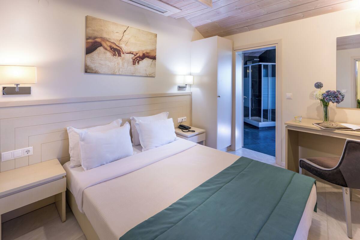 A hotel room with a bed, bedside table, desk, chair, and a painting on the wall.
