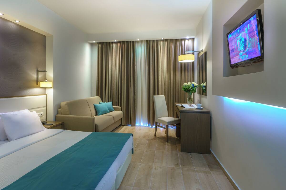 A hotel room with a bed, a sofa, a desk, a chair, a TV, and curtains.