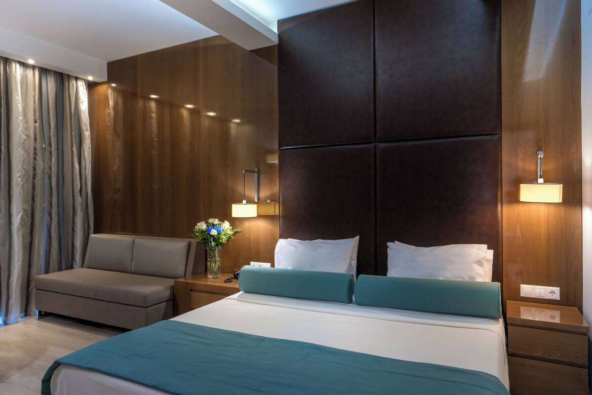 A modern hotel room with a large bed, a sofa, and a nightstand with a lamp.
