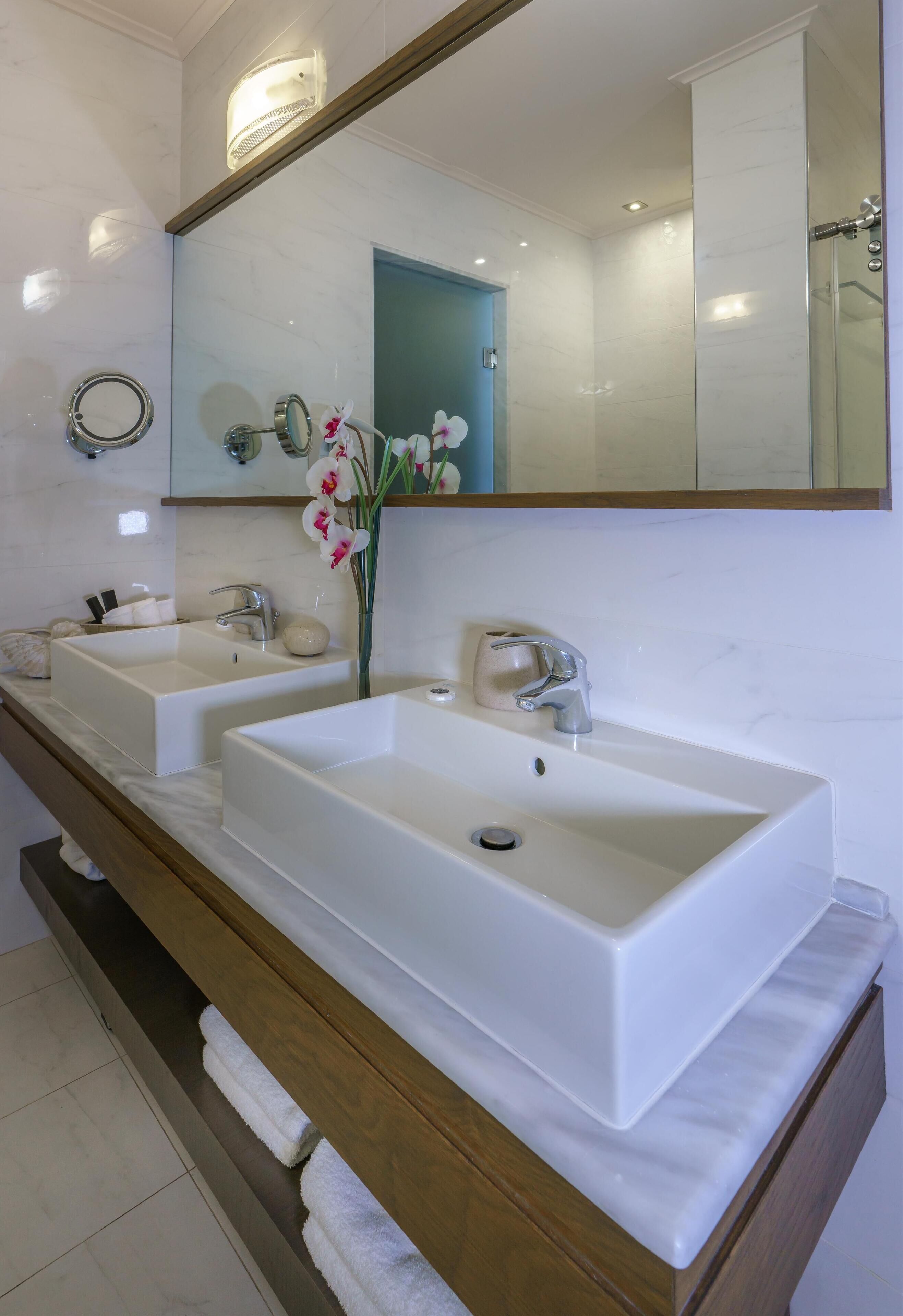 Un baño con dos lavamanos blancos, un mueble de tocador de madera, un espejo y un florero con flores.