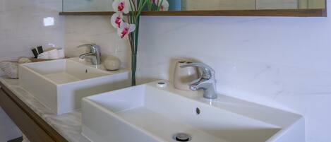 Un baño con dos lavamanos blancos, un mueble de tocador de madera, un espejo y un florero con flores.