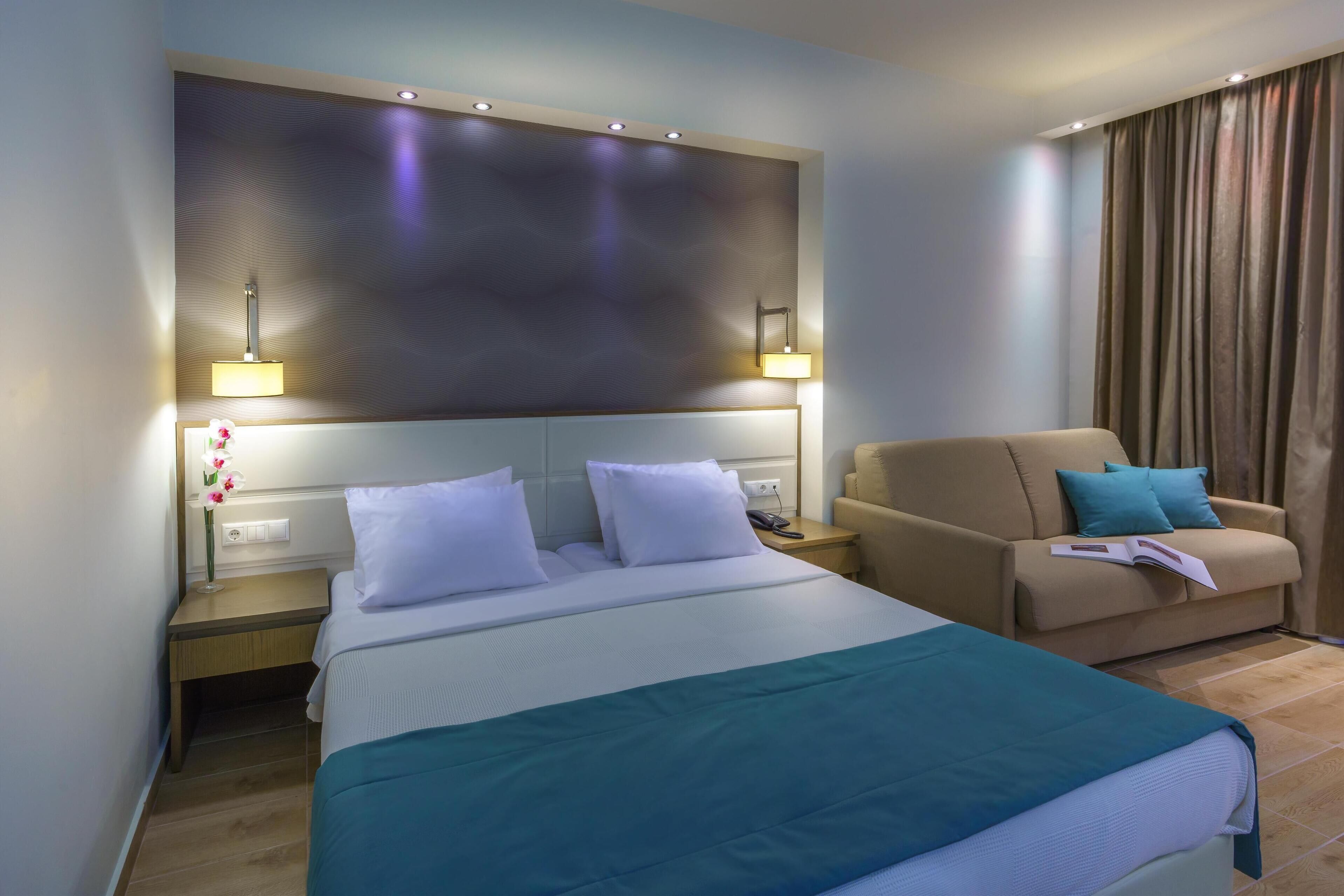 A hotel room with a large bed, a sofa, a nightstand with a lamp, and a wall-mounted light fixture.