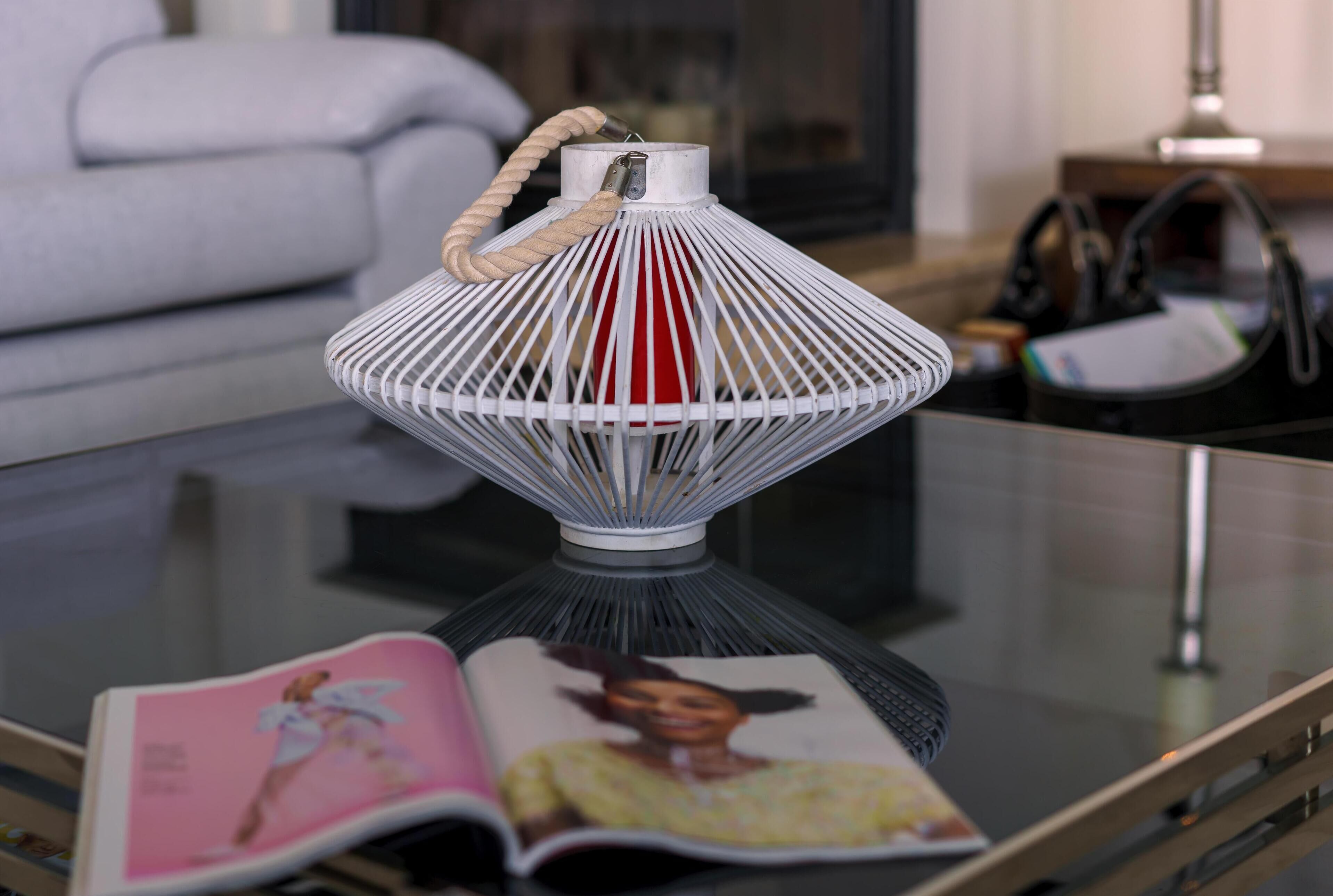 Een witte, geweven lantaarn met touwhal op een glazen tafel, naast een open tijdschrift.