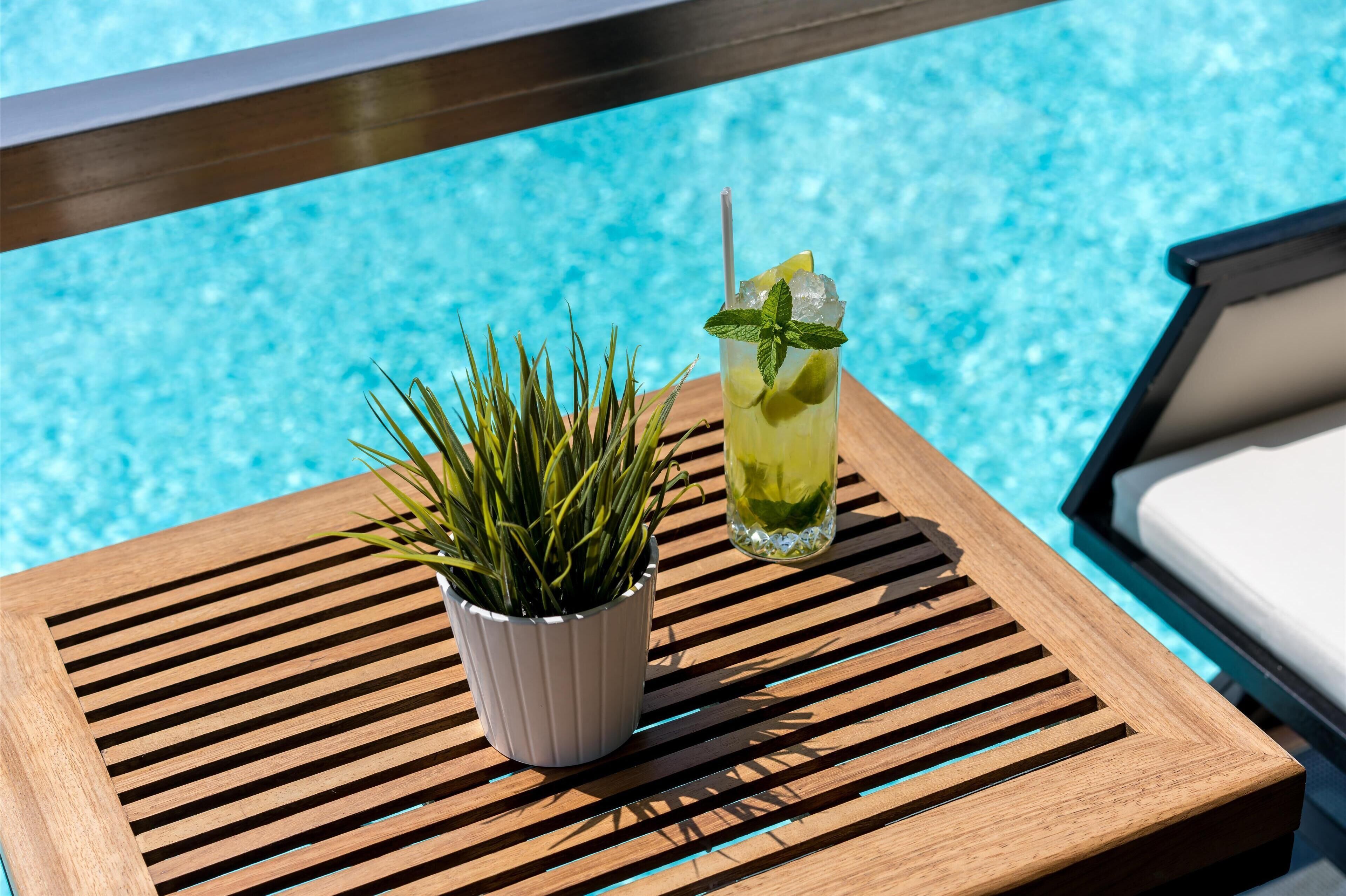 Un tavolo in legno con una pianta in vaso, un bicchiere di bevanda fresca con cannucce e guarnizione di menta, vicino a una piscina.