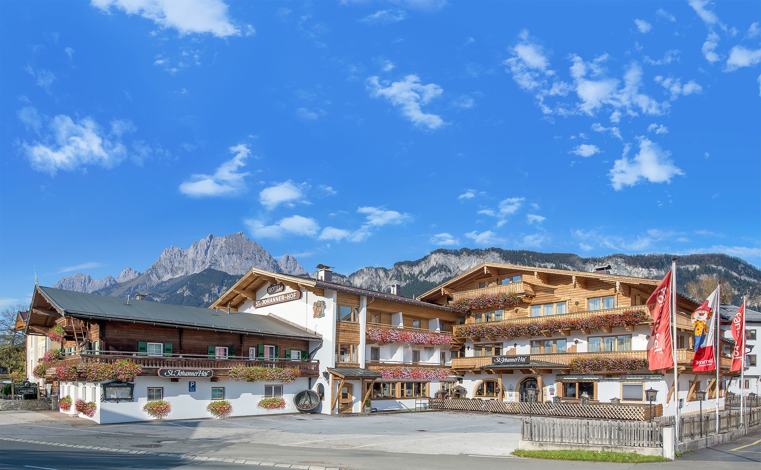 Ein traditionelles Holzhaus mit Balkonen, die mit Blumen geschmückt sind, einem Schild mit der Aufschrift "St. Johannnerhof" und einem Berg im Hintergrund.