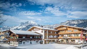 A hotel with multiple wooden balconies, a sign reading "Hotel St. Johannner-Hof," and snow-covered mountains in the background. - Familotel St. Johanner Hof (Sankt Johann in Tirol)