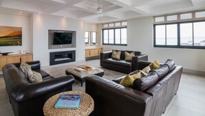 A spacious living room with leather sofas, a flat-screen TV, a fireplace, and large windows offering a view of the sea. - The Gallery Studios (Hermanus)