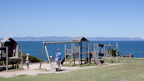 A playground with a swing set and climbing structures near a body of water. - The Gallery Studios (Hermanus)