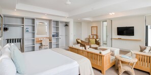 Habitación de hotel con una cama grande, una cuna de madera, una zona de estar con una mesita, un televisor y un armario con estantes incorporados.