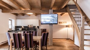 A modern kitchen with white cabinets, a wooden dining table with striped chairs, and a TV mounted on a wooden beam. - A CASA Smaragd (Soelden)