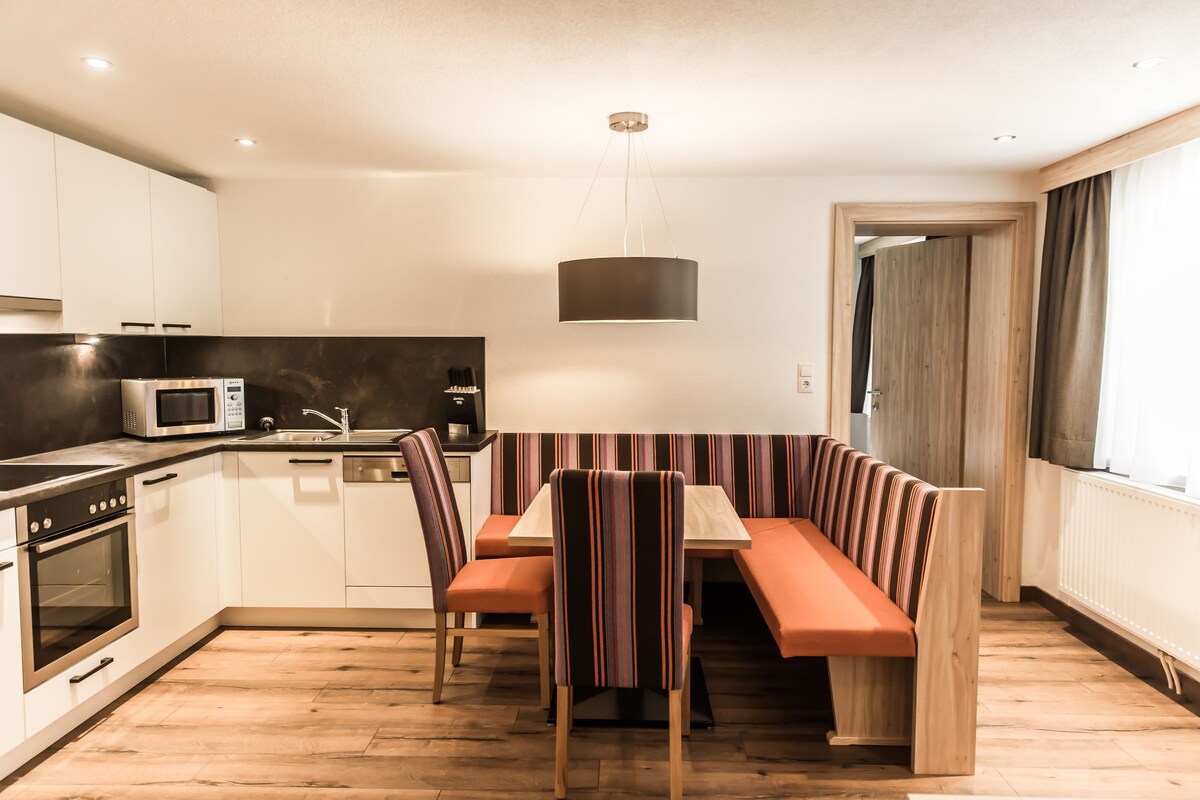 A modern kitchen with white cabinets, a microwave, and a sink. There is a wooden dining table with striped chairs and a bench.