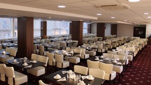 Un amplio salón de banquetes con numerosas mesas dispuestas para comer, techos altos con iluminación empotrada y grandes ventanas que dejan entrar luz natural.