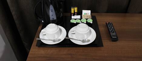 A hotel room with a wooden table set for breakfast, including two white cups, spoons, a black coffee maker, tea packets, a remote control, and a card.