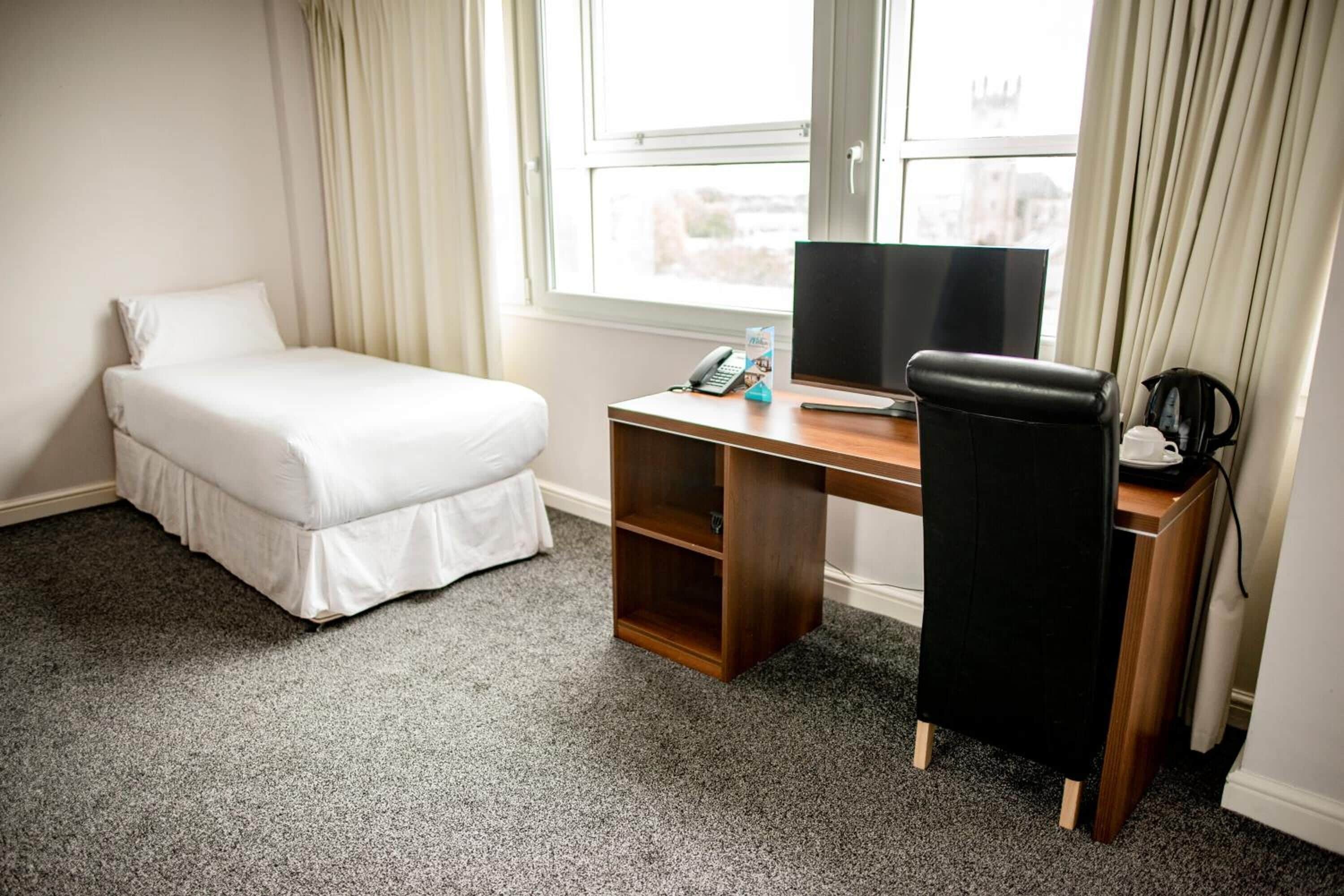 A hotel room with a bed, a desk with a computer, a chair, and a telephone.