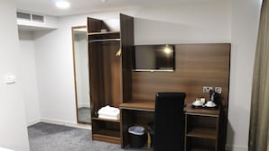 Habitación de hotel con un escritorio de madera, una silla negra, un televisor montado en la pared, un armario y un pequeño cubo de basura.