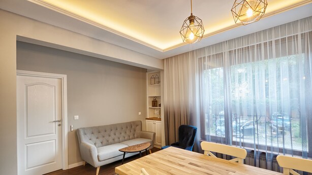 Un salon moderne avec un canapé, une table à manger et une chaise. On y trouve une fenêtre avec des rideaux, un luminaire au plafond et une porte.