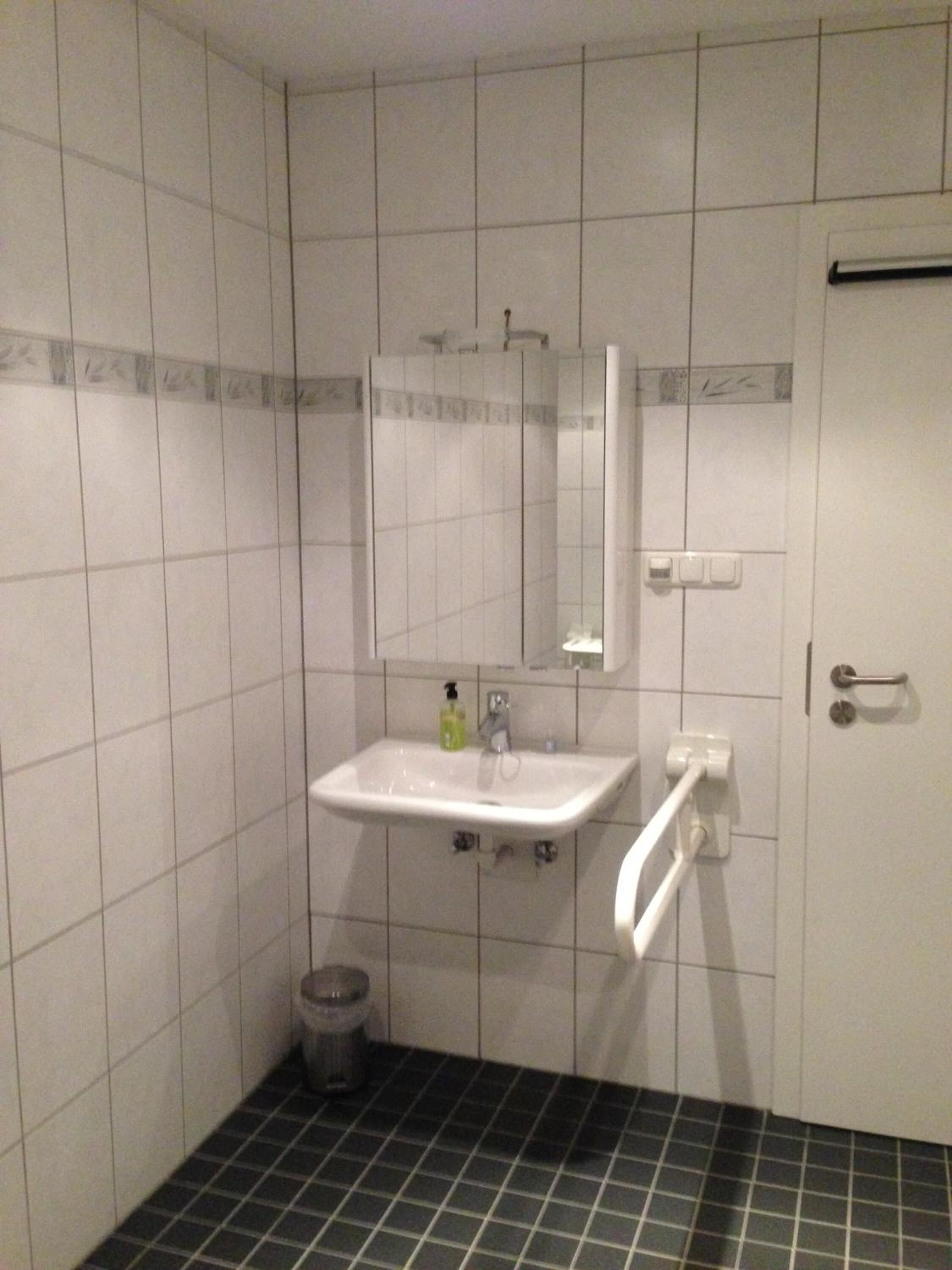 Un baño con azulejos blancos, un lavamanos, un espejo, un perchero para toallas y una puerta.