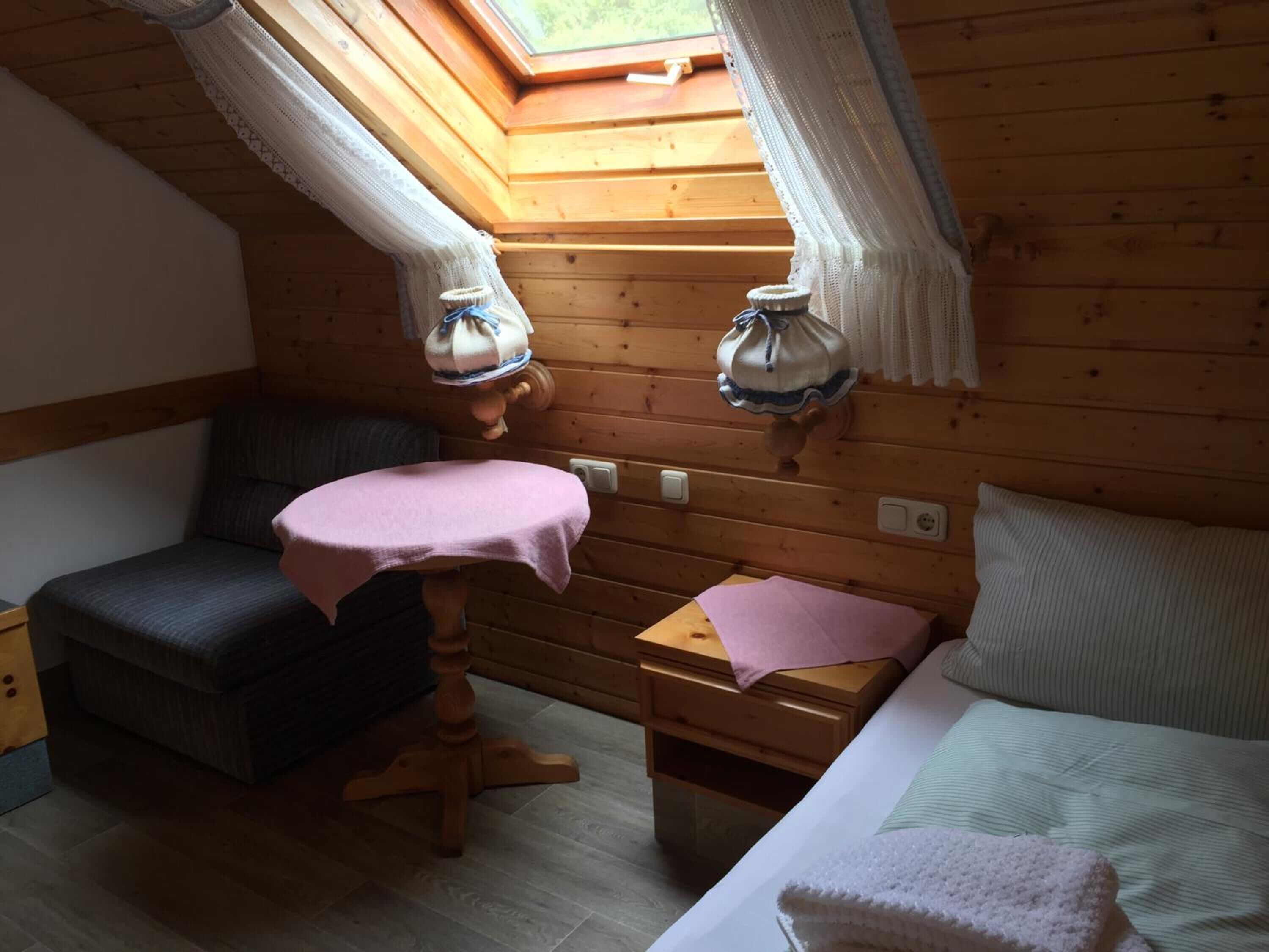 A cozy cabin room with a wooden interior, a skylight, a small table with a pink cloth, a bed with a white pillow, and a nightstand with a pink cloth.